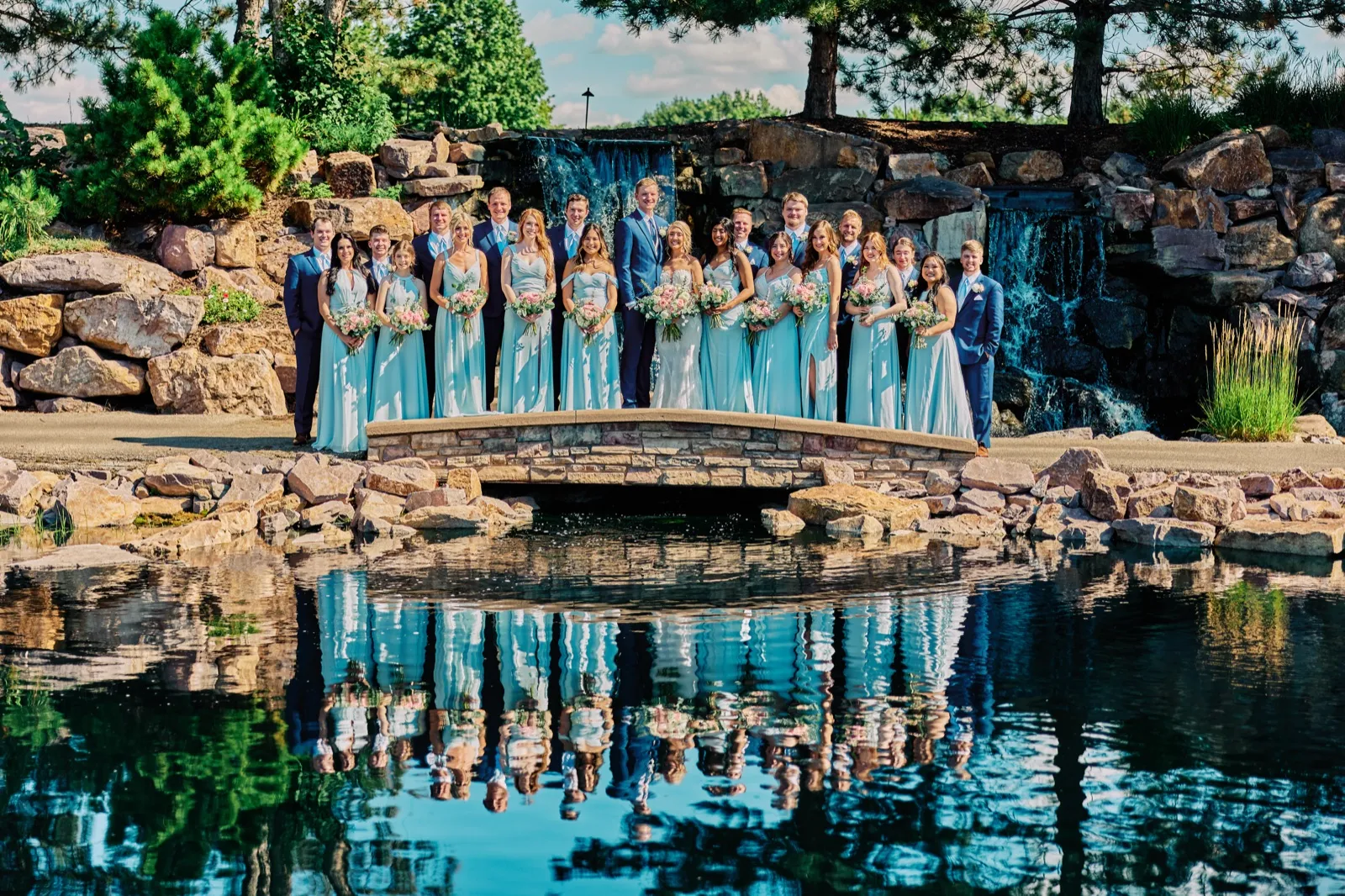 The full bridal party stands on a stone bridge in front of a waterfall, with their reflection mirrored in the pond below. — Tim Larsen Photography, Twin Cities MN