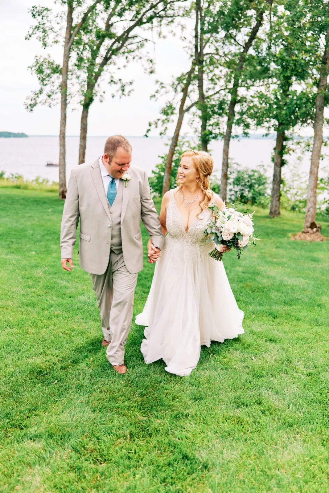 The bride and groom walk hand in hand across a grassy lawn beneath tall trees with Gull Lake visible behind them. — Tim Larsen Photography, Brainerd Lakes MN