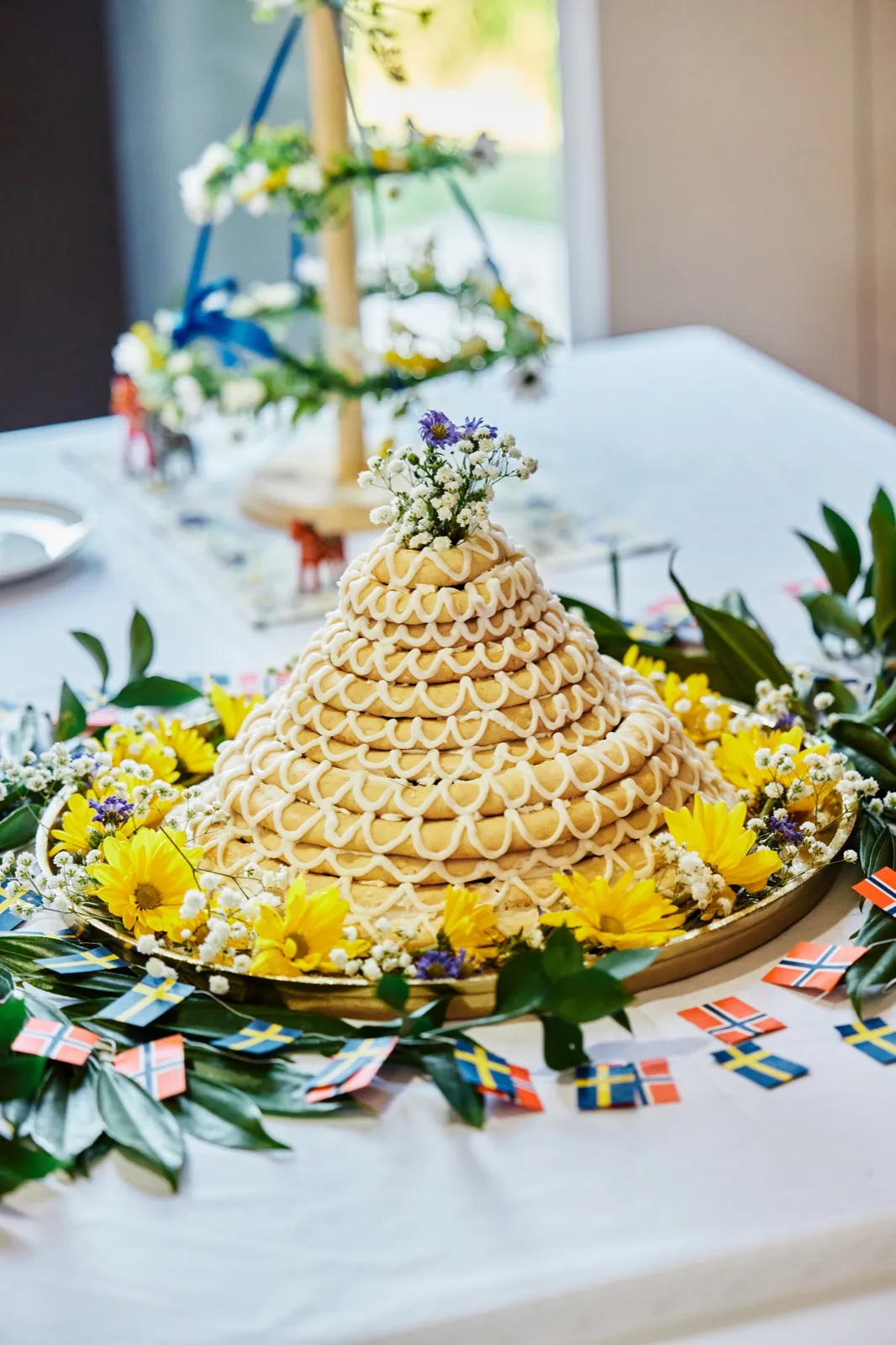A tiered Scandinavian kransekake topped with wildflowers sits on a gold platter encircled by yellow daisies, baby's breath, and small Swedish and Norwegian flag garlands. — Tim Larsen Photography, Minnesota