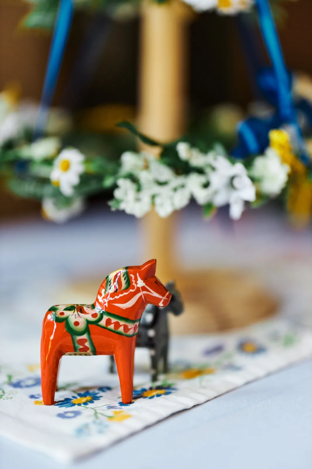 A small red painted Swedish Dala horse sits on an embroidered linen with a daisy flower crown and blue ribbon blurred in the background. — Tim Larsen Photography, Minnesota