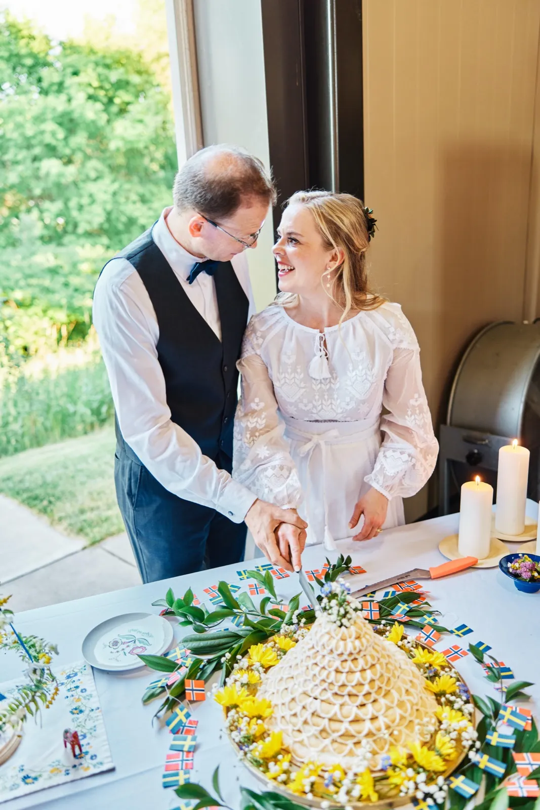 The bride and groom share a smiling glance as they cut into a tall Scandinavian kransekake cake surrounded by Swedish and Norwegian flags, sunflowers, and pillar candles. — Tim Larsen Photography, Minnesota