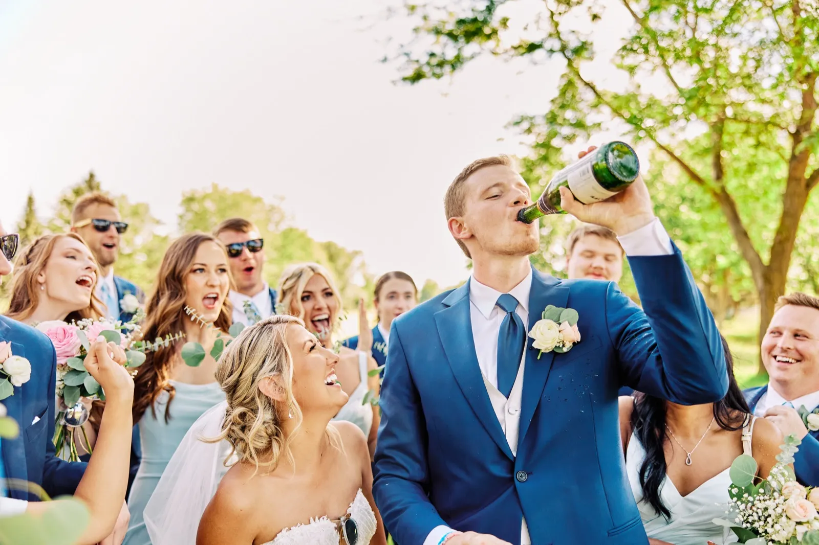 The groom tips a champagne bottle back to drink while the bride and bridal party laugh and cheer around him on a sunlit lawn. — Tim Larsen Photography, Twin Cities MN