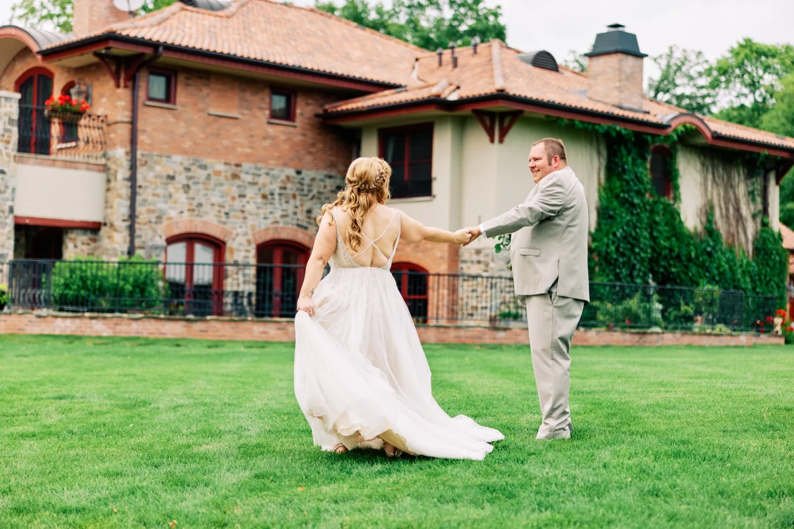 The bride lifts the skirt of her gown as she holds the groom's hand on a wide green lawn in front of a stone-and-brick estate covered in ivy. — Tim Larsen Photography, Brainerd Lakes MN