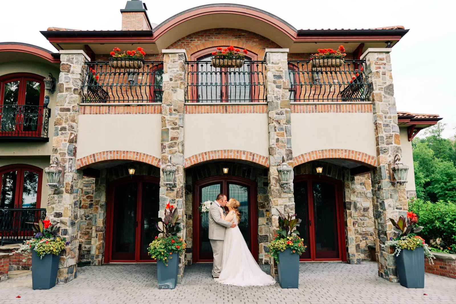 The bride and groom share a kiss in front of a grand stone mansion with arched balconies, red doors, and flower-filled planters. — Tim Larsen Photography, Brainerd Lakes MN