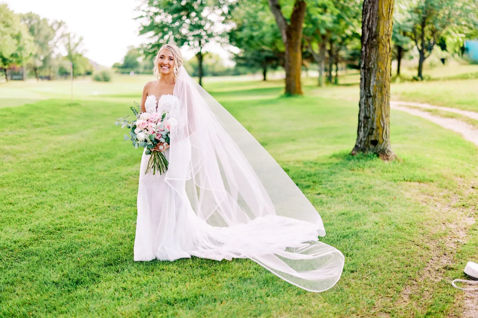 The bride stands smiling on a sunlit golf course lawn holding a pink and white bouquet as her cathedral-length veil trails across the grass. — Tim Larsen Photography, Twin Cities MN