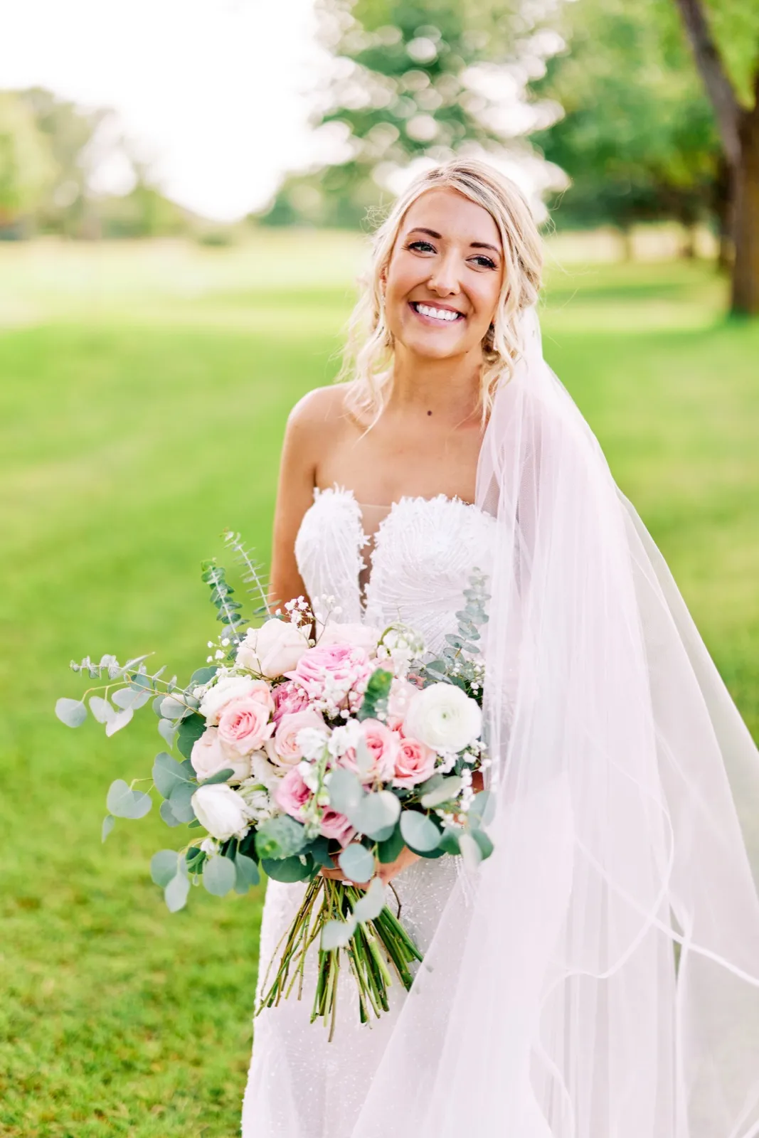 The bride smiles broadly while holding a pink and white rose bouquet, her long veil trailing to one side on a sunlit lawn. — Tim Larsen Photography, Twin Cities MN