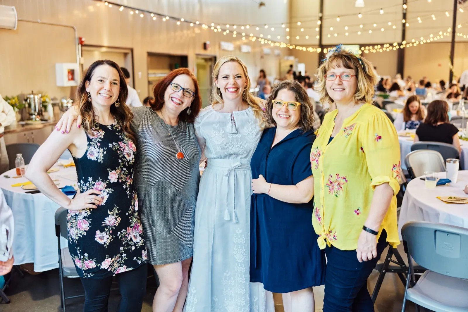 The bride poses arm-in-arm with four women guests beneath string lights in the reception hall, all smiling toward the camera. — Tim Larsen Photography, Minnesota