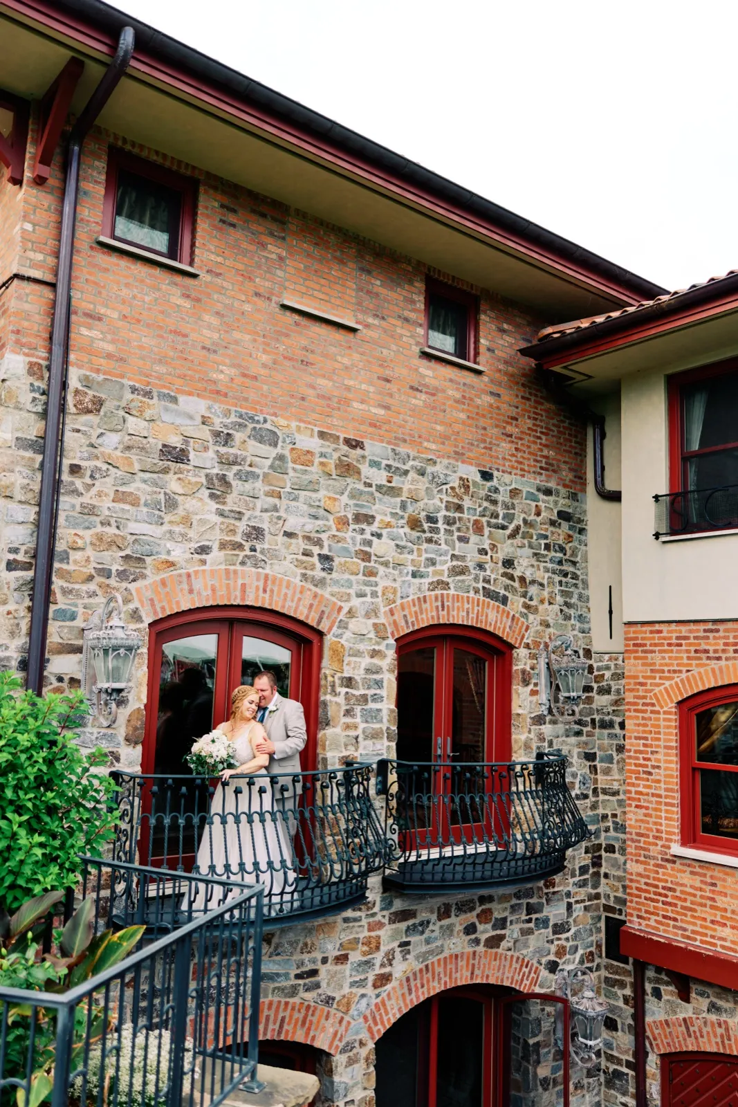The bride and groom share a quiet embrace on a small wrought-iron balcony surrounded by brick and stone walls with red-trimmed arched doors. — Tim Larsen Photography, Brainerd Lakes MN