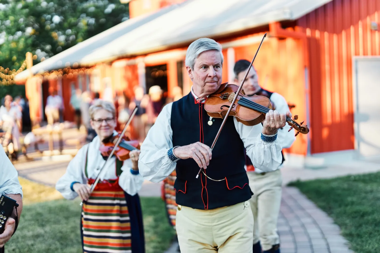 A violinist in traditional Scandinavian folk dress plays as he walks past a red barn, with a second fiddler in a striped folk skirt following behind in evening light. — Tim Larsen Photography, Minnesota