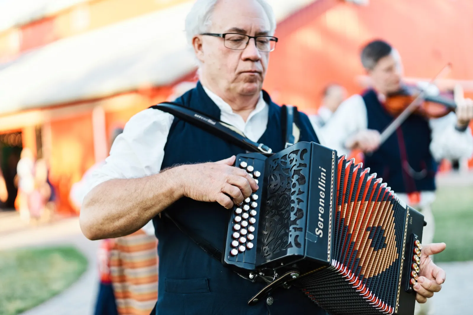An accordion player in a black vest concentrates on his Serenellini accordion while a violinist plays in the background outside a barn in warm evening light. — Tim Larsen Photography, Minnesota