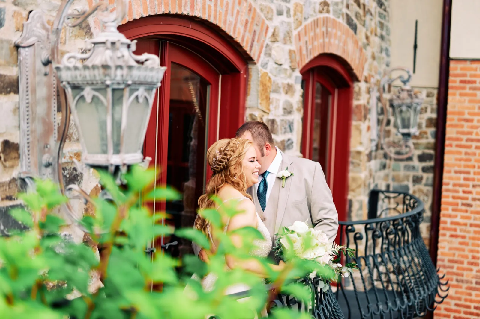 The bride leans into the groom and laughs while holding a cream-and-blush bouquet, framed by green foliage and an ornate iron lantern outside a stone building with red arched doors. — Tim Larsen Photography, Brainerd Lakes MN