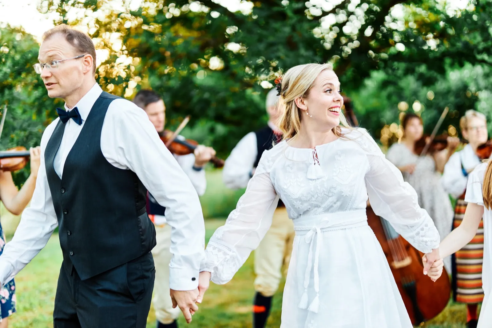 The bride laughs joyfully while holding the groom's hand as folk musicians playing violins and a cello surround the couple on a sunlit lawn. — Tim Larsen Photography, Minnesota