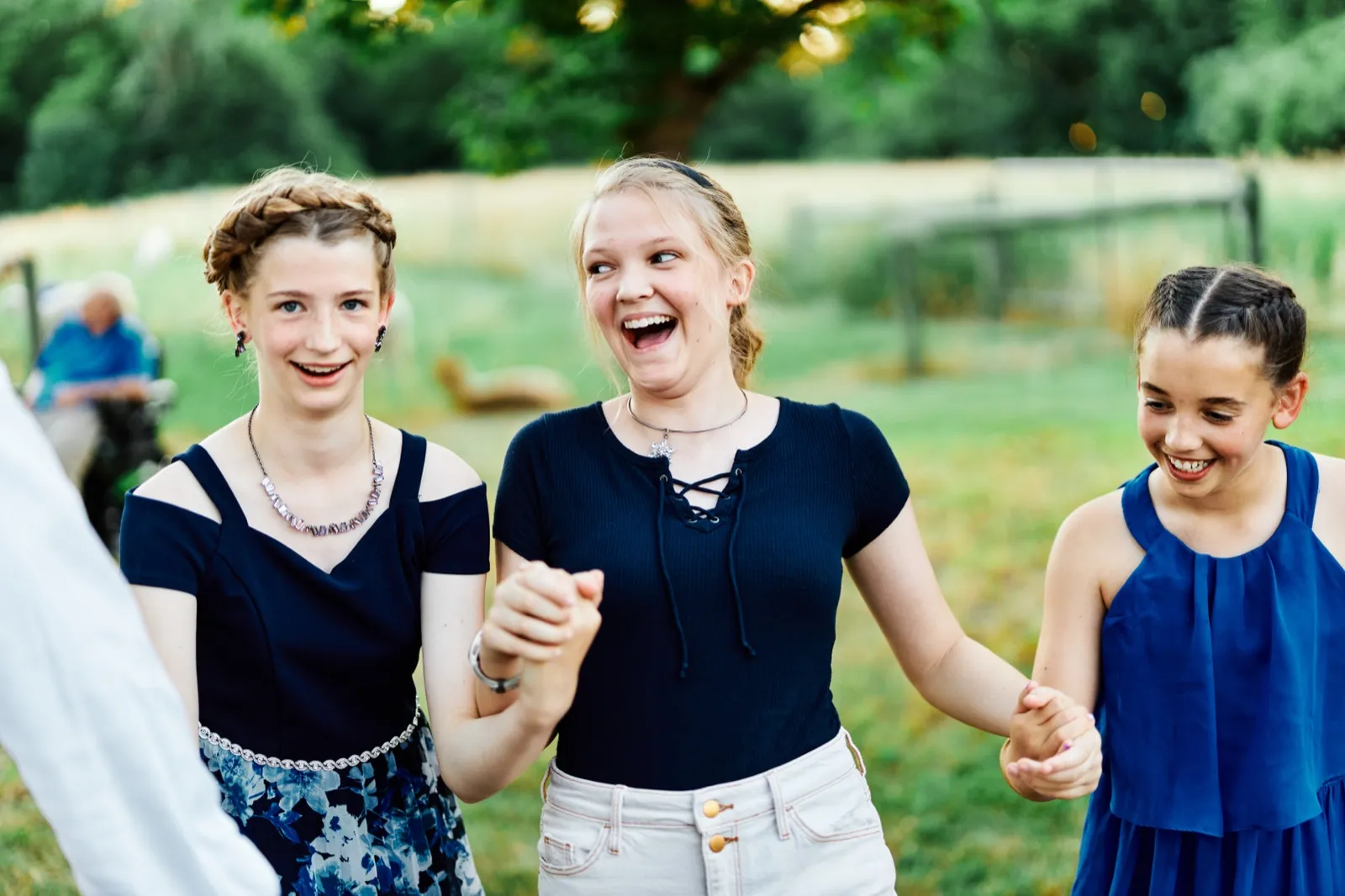 Three young guests in dark blue dresses laugh together while holding hands in a sunlit farm field at evening. — Tim Larsen Photography, Minnesota