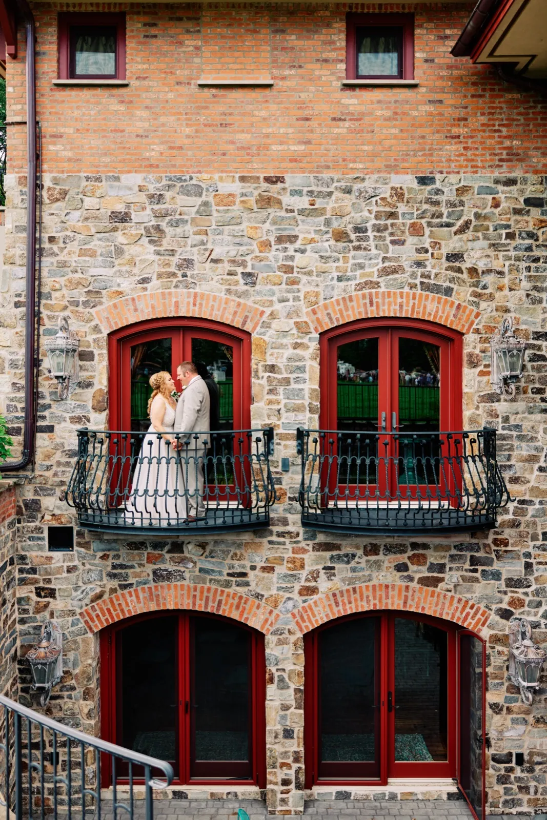 The bride and groom stand together on a small wrought-iron balcony framed by red French doors set into a stone-and-brick facade. — Tim Larsen Photography, Brainerd Lakes MN