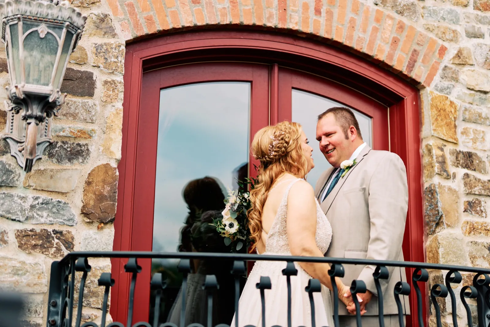The bride and groom stand close together on a wrought-iron balcony beneath a red arched doorway set into a stone facade, smiling at each other as she holds a white bouquet. — Tim Larsen Photography, Brainerd Lakes MN