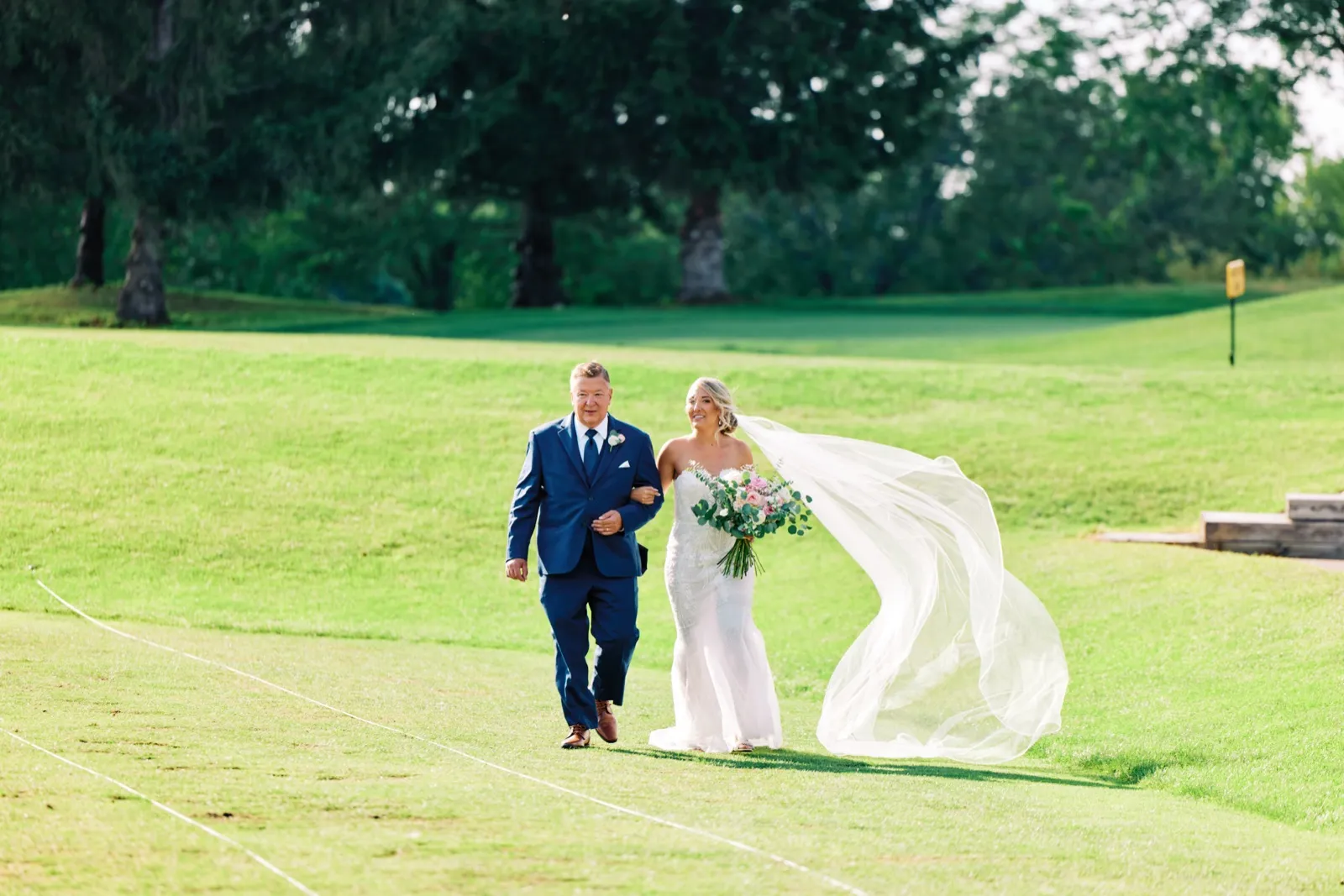 The bride walks across a sunlit golf course fairway on her father's arm, her long veil sweeping sideways in the wind as she smiles ahead. — Tim Larsen Photography, Twin Cities MN