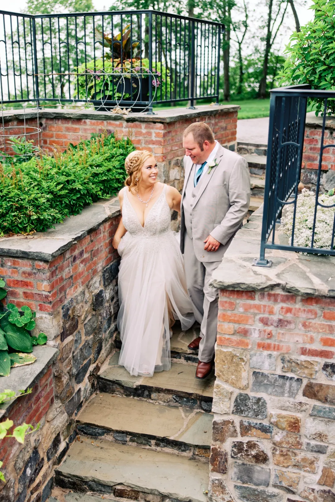 The bride and groom share a look while standing on stone steps surrounded by brick walls and iron railings in a lakeside garden. — Tim Larsen Photography, Brainerd Lakes MN