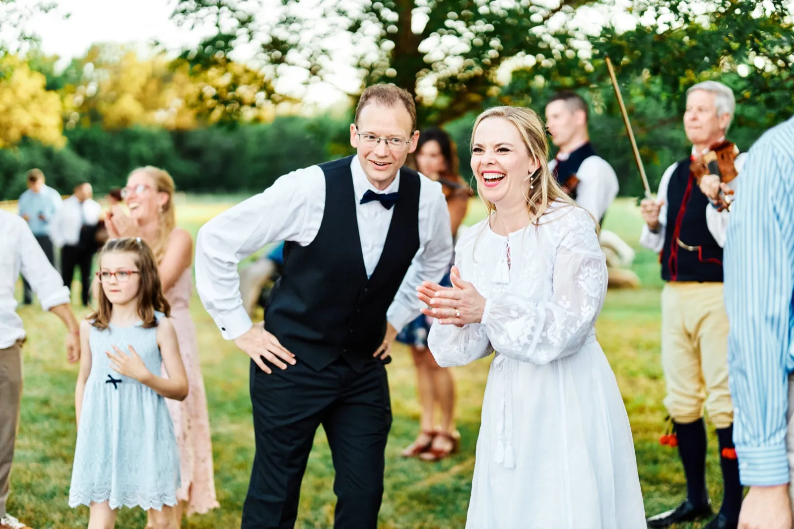 The bride claps and laughs beside the groom on a grassy lawn while folk musicians in traditional vests play violins behind them. — Tim Larsen Photography, Minnesota