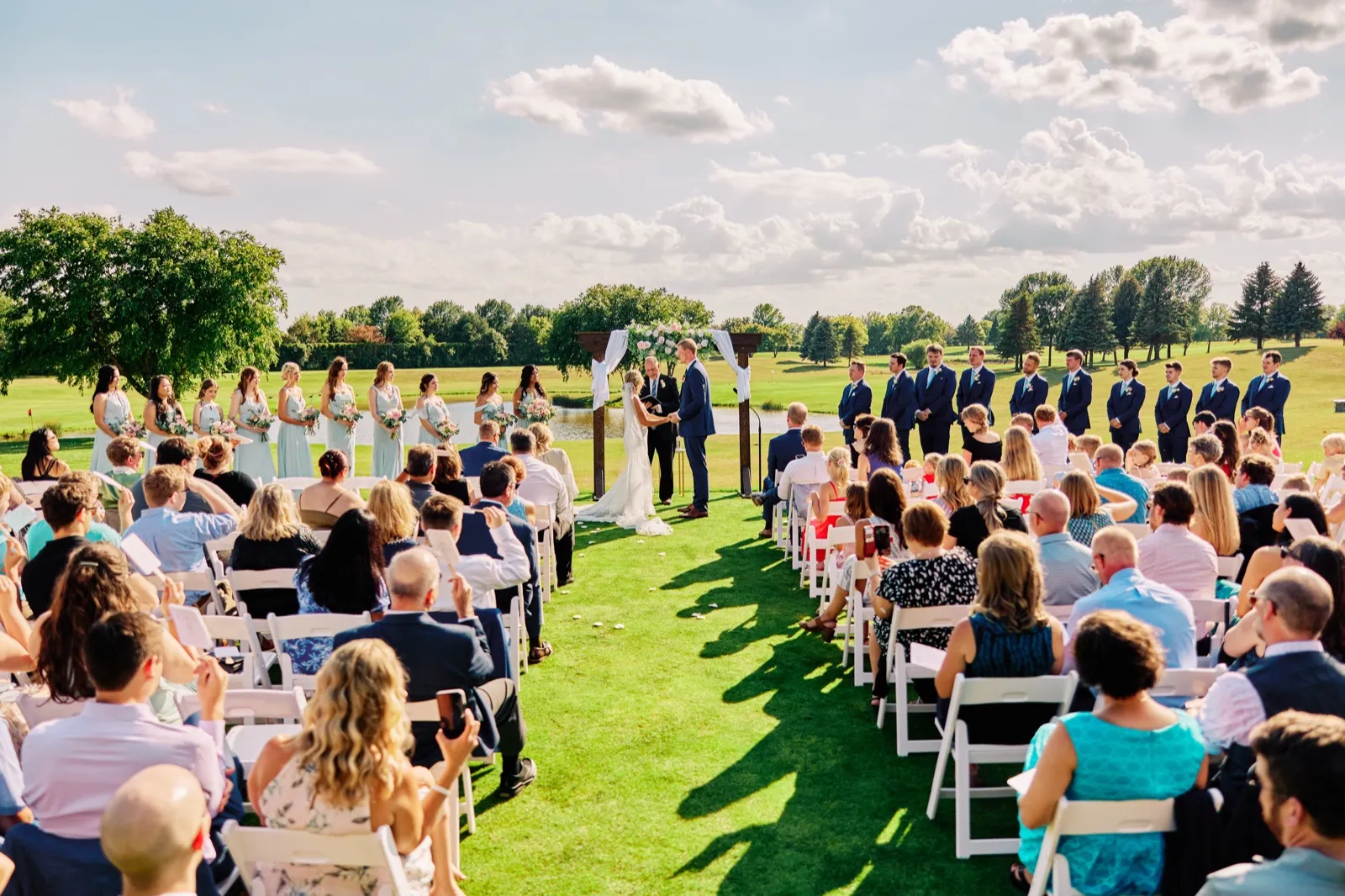 A wide view down the aisle of an outdoor ceremony at a golf course, with the couple beneath a floral arch, bridal party flanking them, and seated guests on either side under a partly cloudy sky. — Tim Larsen Photography, Twin Cities MN