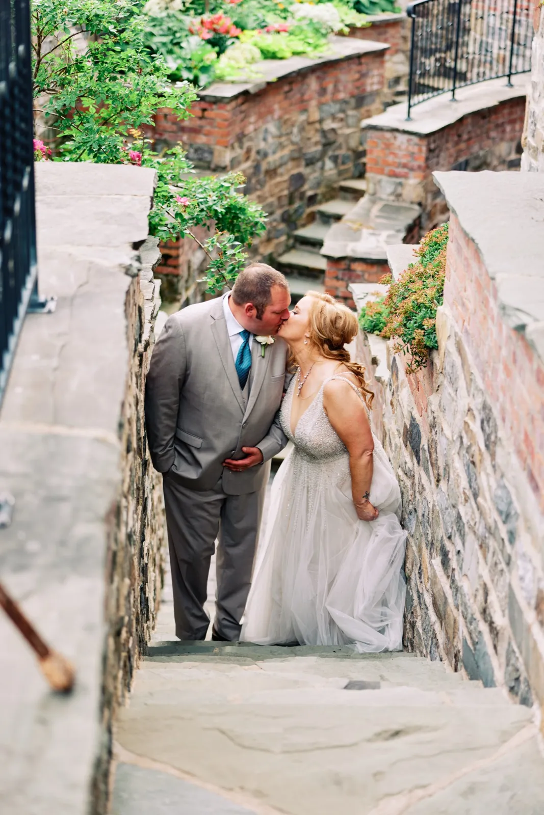 The couple shares a kiss on a stone stairway flanked by brick walls and overflowing greenery, photographed from above. — Tim Larsen Photography, Brainerd Lakes MN