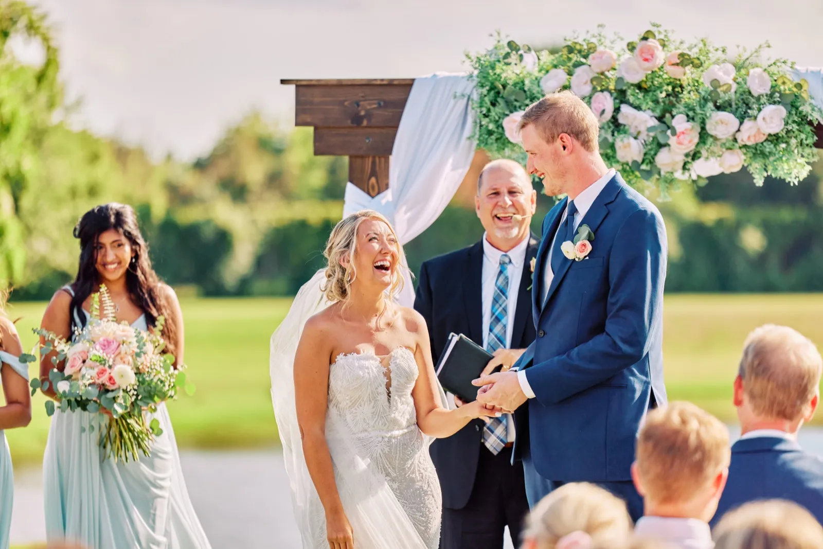 The bride throws her head back laughing as she holds the groom's hand at the ceremony arch, with the officiant smiling beside them and a bridesmaid watching from the left. — Tim Larsen Photography, Twin Cities MN