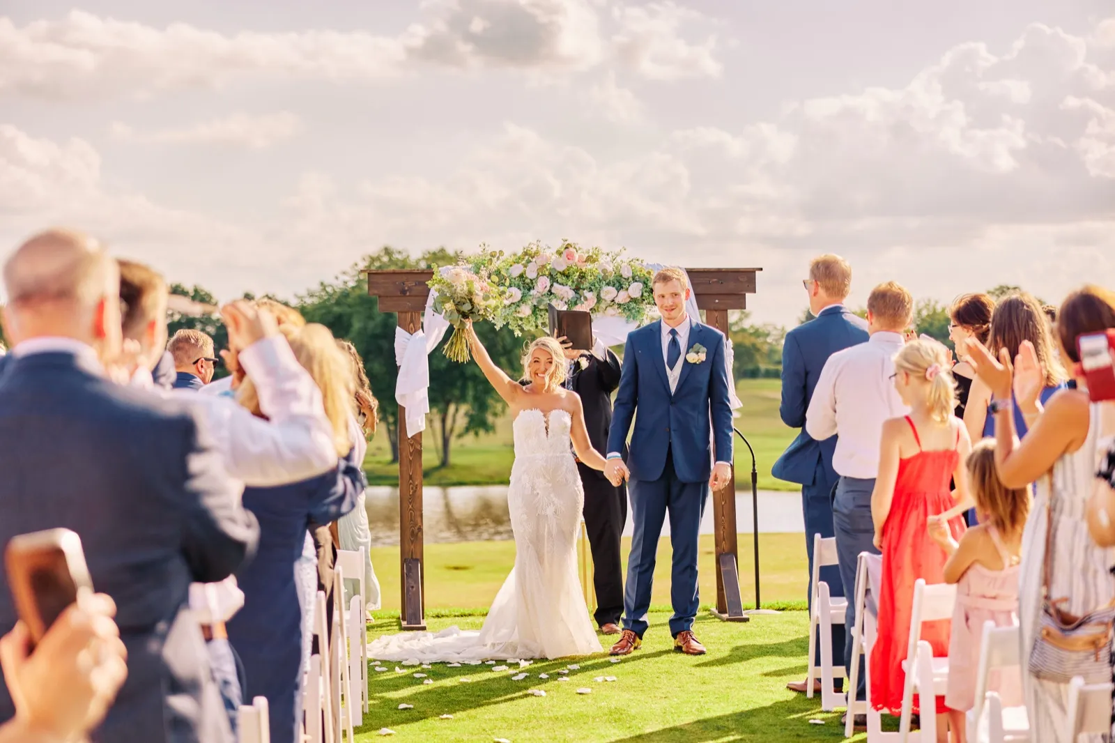 The bride raises her bouquet overhead and smiles beside the groom under a floral ceremony arch as guests applaud from both sides of the aisle. — Tim Larsen Photography, Twin Cities MN