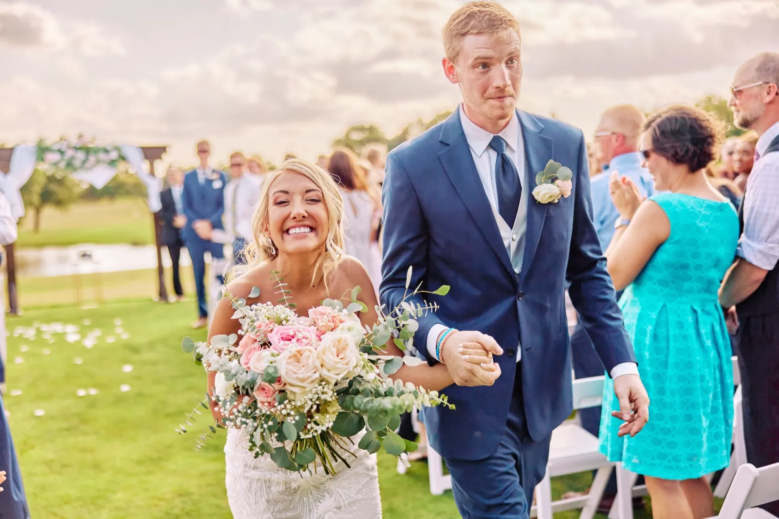 The bride beams with a wide smile as she walks down the aisle holding her bouquet, her groom beside her holding her hand, with guests applauding on either side. — Tim Larsen Photography, Twin Cities MN