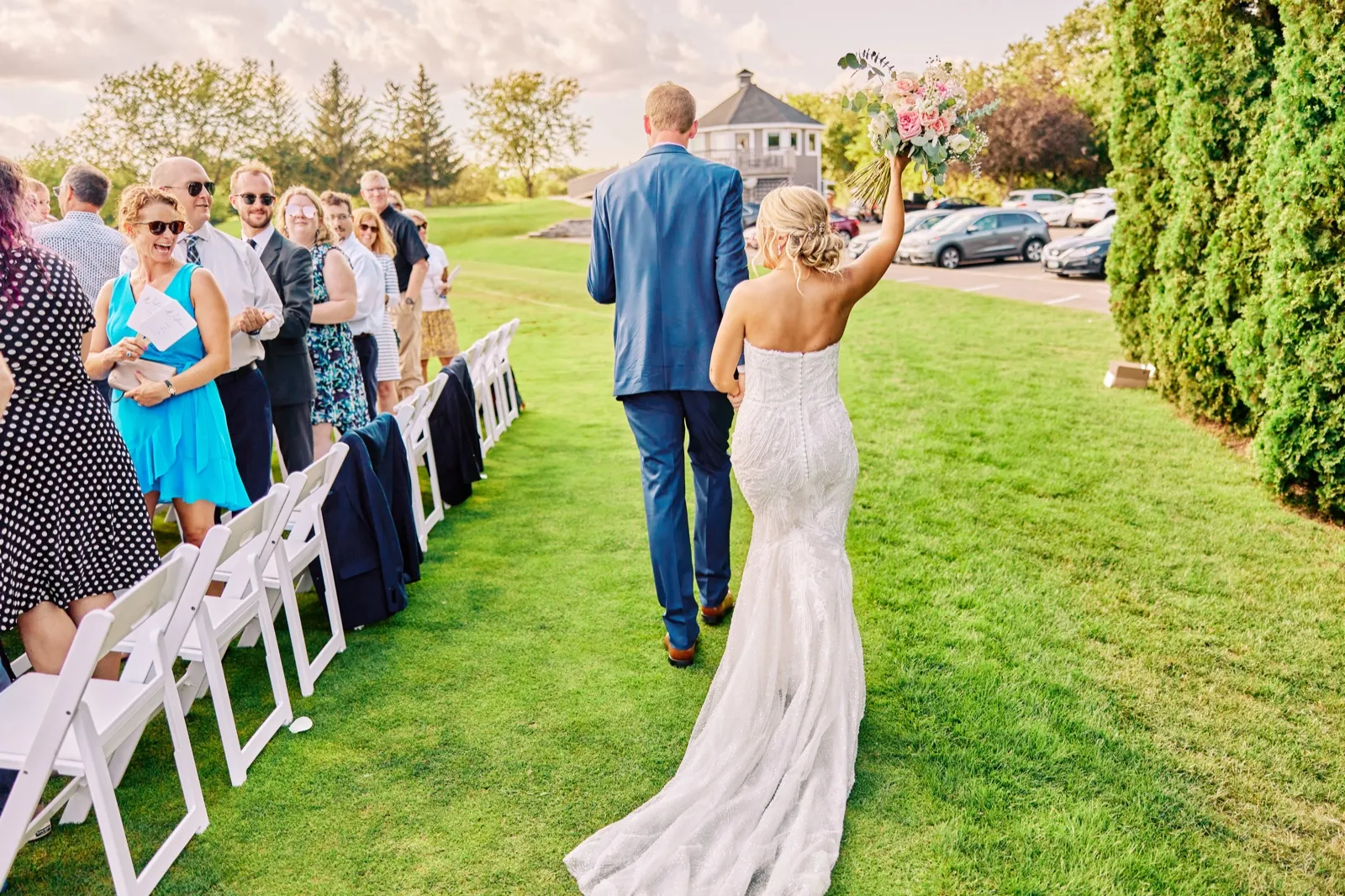 The bride raises her bouquet overhead in celebration as she walks back down the aisle with the groom, cheering guests visible along the row of white chairs. — Tim Larsen Photography, Twin Cities MN