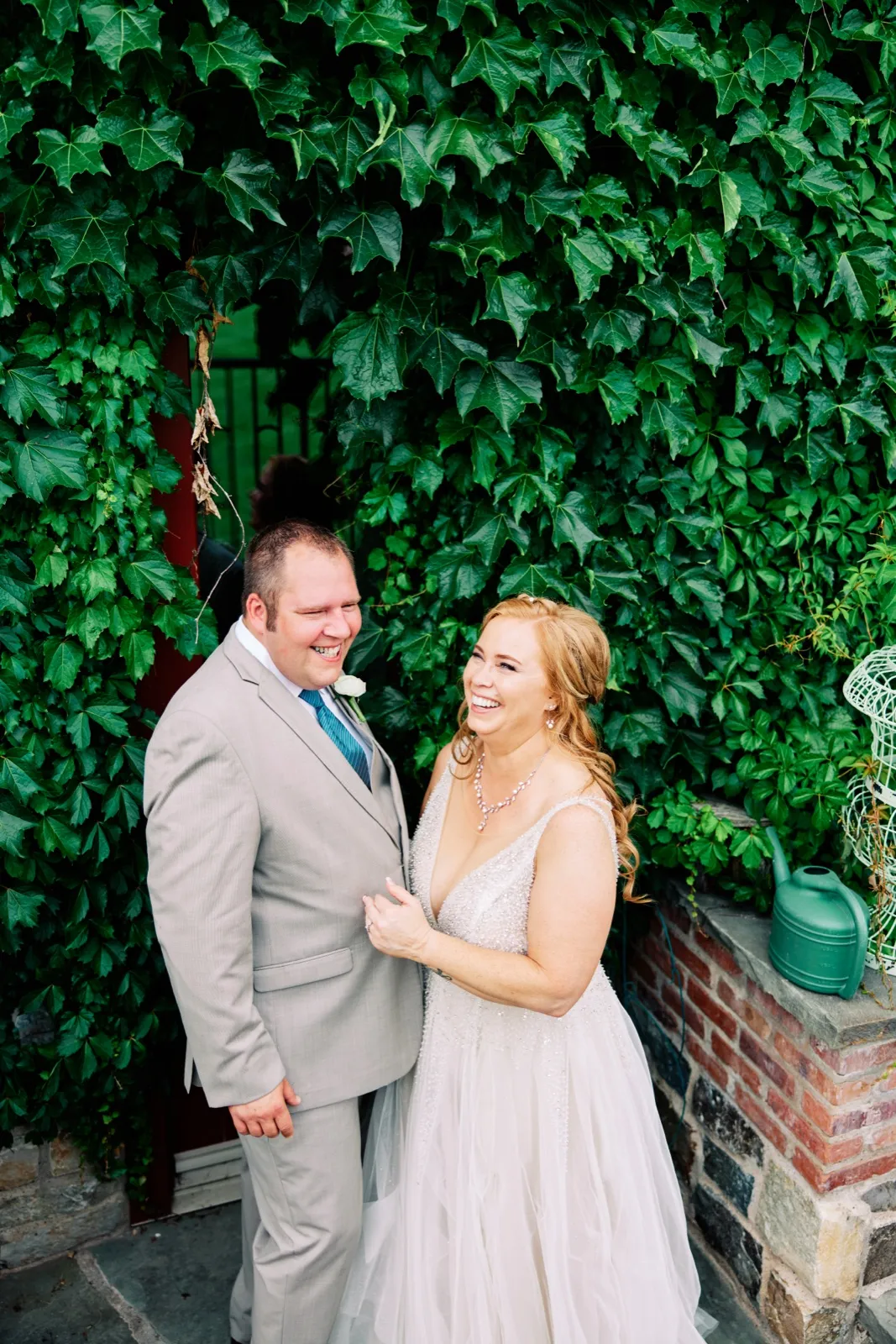 The bride and groom laugh together while standing in front of a tall ivy-covered wall, with a brick ledge and watering can to one side. — Tim Larsen Photography, Brainerd Lakes MN