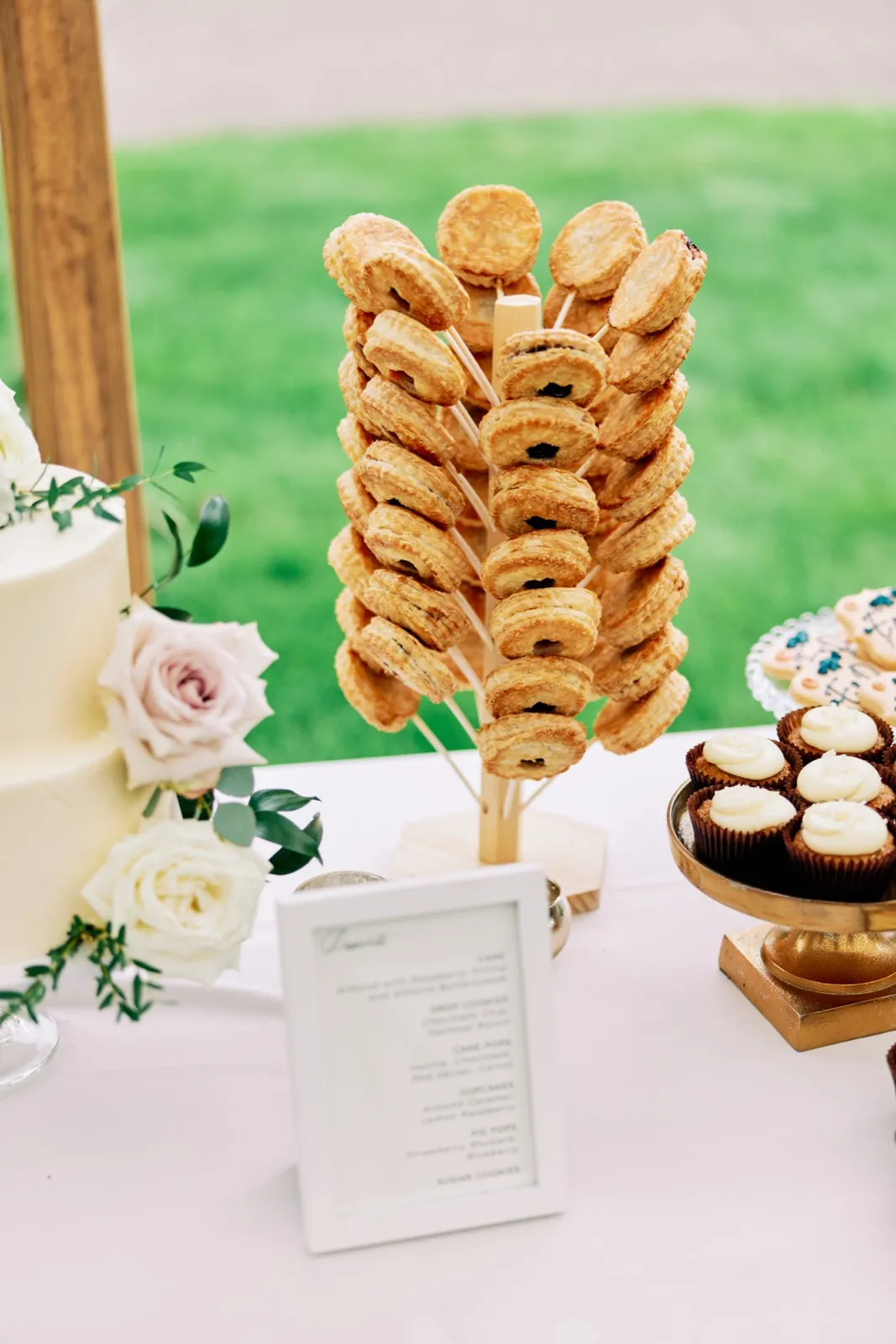 A dessert table under a tent featuring a tiered white cake with roses, a tall wooden tower of pie pops, a gold stand of cupcakes, and a framed dessert menu. — Tim Larsen Photography, Brainerd Lakes MN