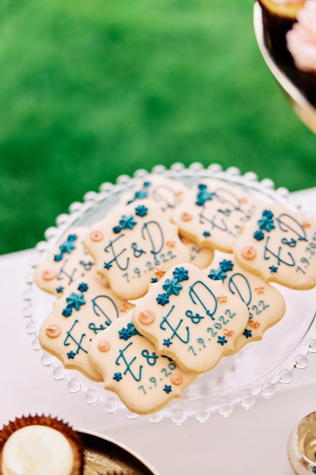Custom monogrammed sugar cookies with the couple's initials and wedding date arranged on a beaded glass cake stand on a grass lawn. — Tim Larsen Photography, Brainerd Lakes MN