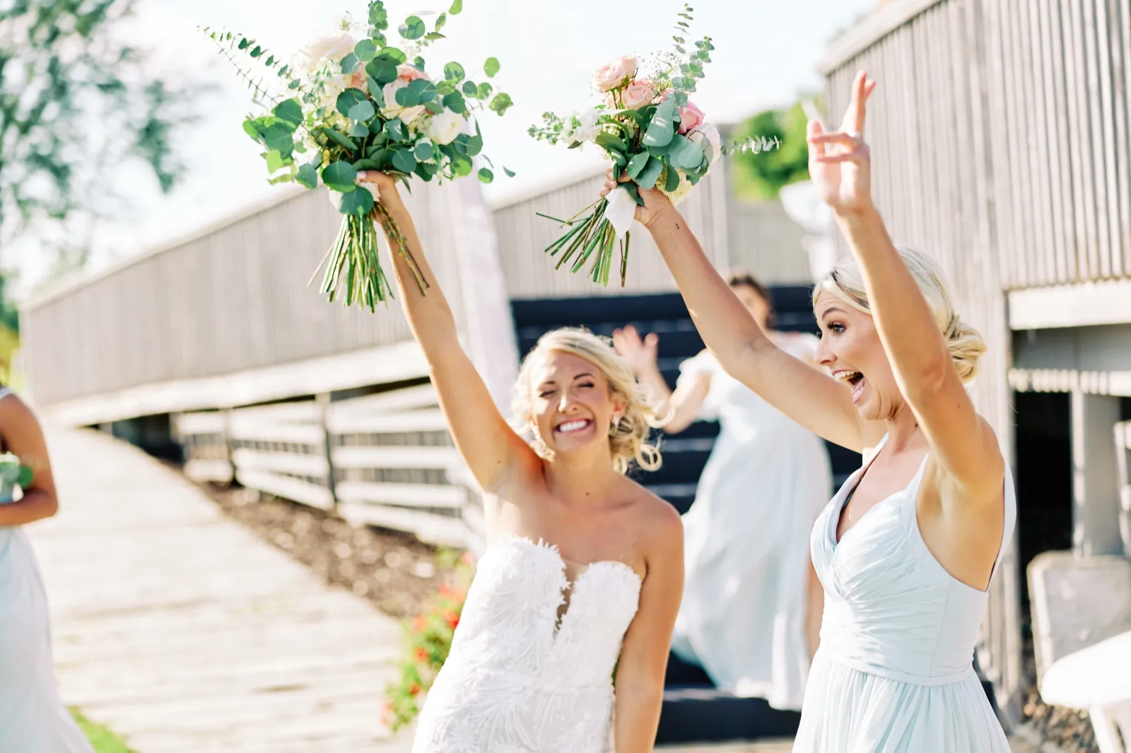 The bride and a bridesmaid raise their bouquets overhead and cheer with open-mouth laughter on a wooden walkway outside the venue. — Tim Larsen Photography, Twin Cities MN