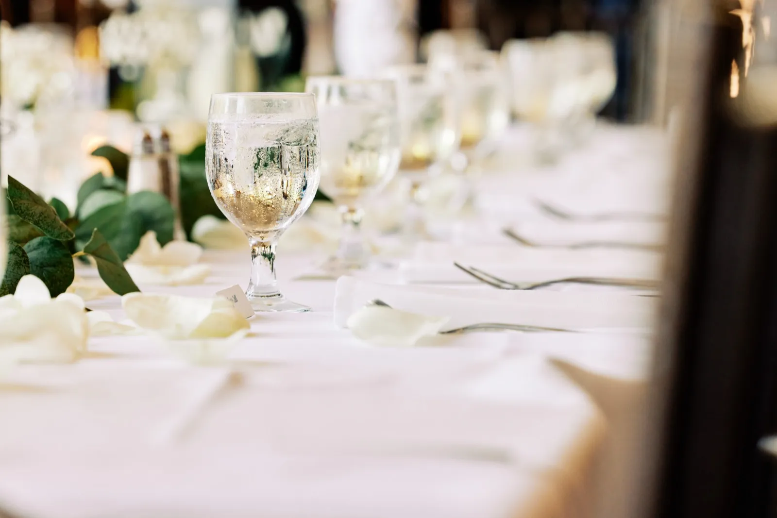 A condensation-beaded water goblet sits on a long white reception table scattered with rose petals and greenery, with more glassware receding into soft focus. — Tim Larsen Photography, Twin Cities MN