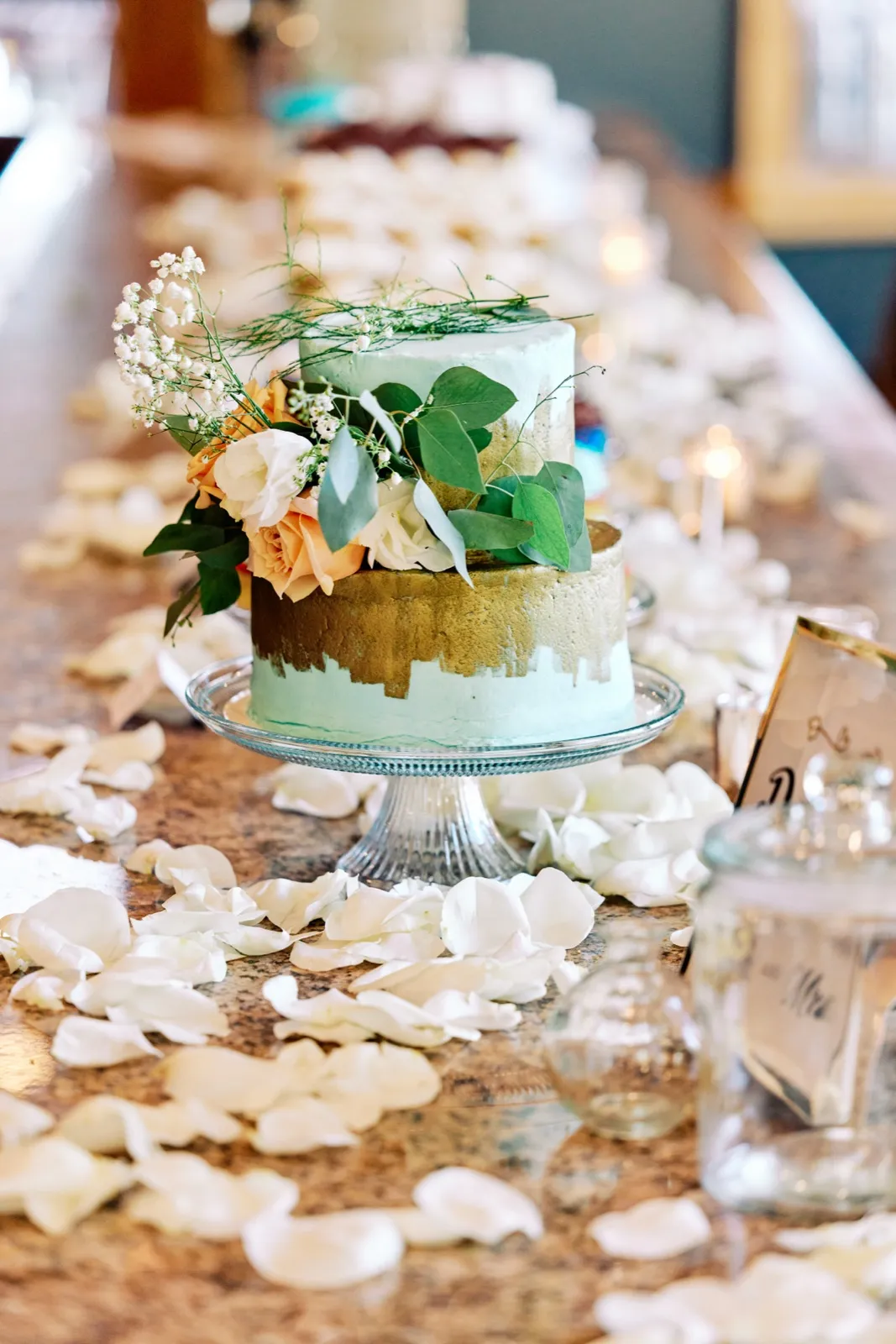 A two-tier mint and gold buttercream wedding cake topped with garden roses and greenery sits on a glass cake stand, surrounded by scattered white rose petals on a granite bar. — Tim Larsen Photography, Twin Cities MN