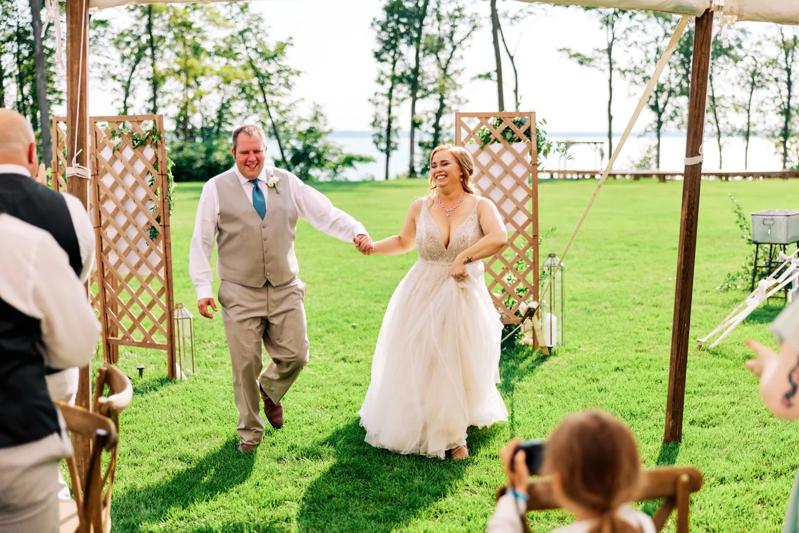 The bride and groom walk hand-in-hand back down the aisle, both laughing, framed by lattice ceremony arches with Gull Lake visible in the background. — Tim Larsen Photography, Brainerd Lakes MN