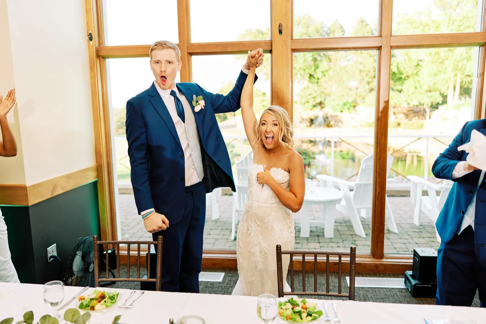 The bride and groom raise their joined hands and shout in celebration as they enter the reception room with the golf course visible through tall windows behind them. — Tim Larsen Photography, Twin Cities MN