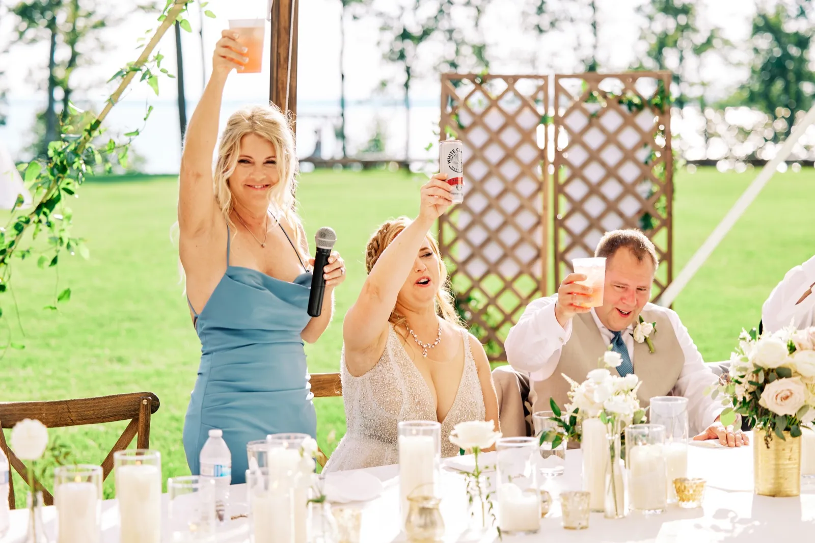 A bridesmaid holding a microphone raises her drink mid-toast as the bride and groom lift their cans and cup in response at a lakeside reception table. — Tim Larsen Photography, Brainerd Lakes MN