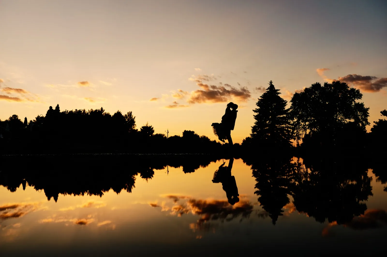 The couple kisses in silhouette against an orange sunset sky, their figures and the surrounding trees mirrored perfectly in the still water of a pond below. — Tim Larsen Photography, Twin Cities MN