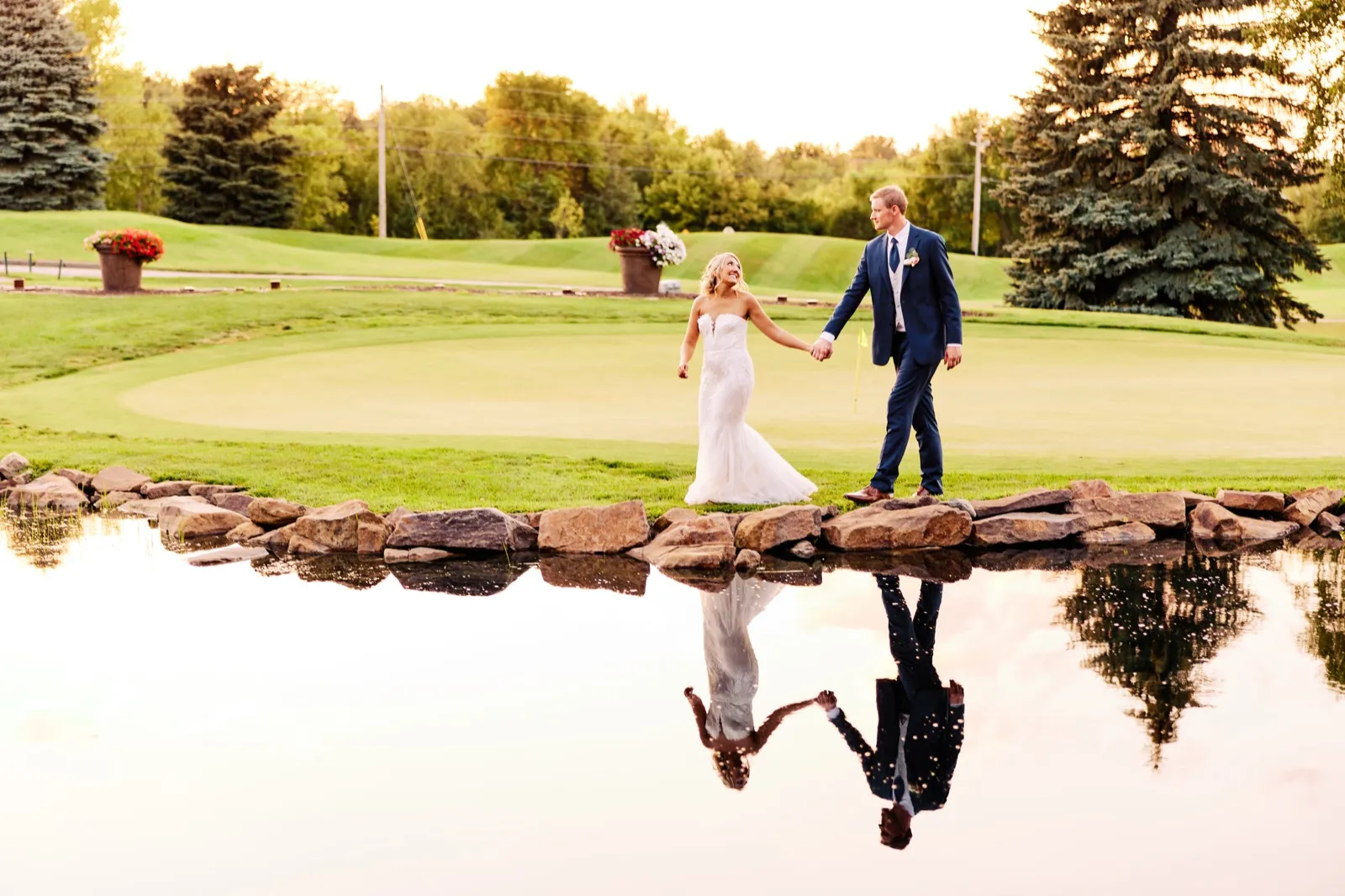 The bride and groom hold hands while walking along a stone-edged pond on a golf course at sunset, their figures mirrored in the still water below. — Tim Larsen Photography, Twin Cities MN