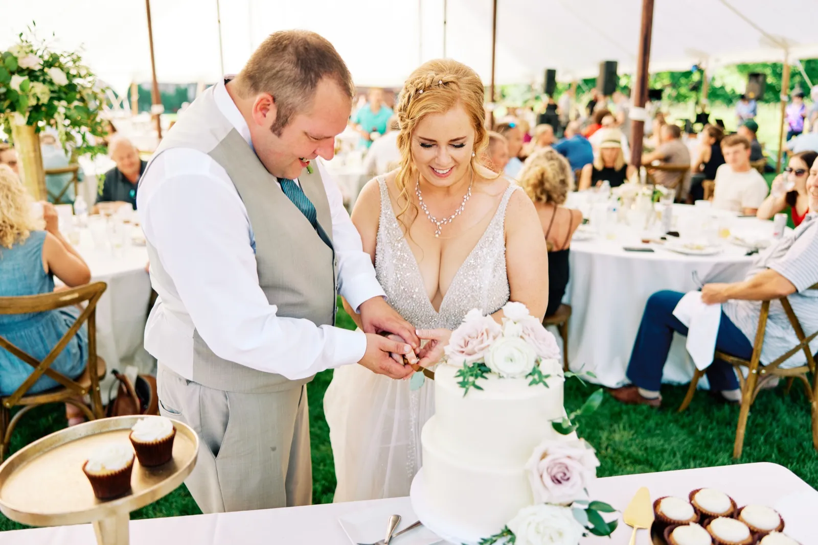 The bride smiles down at a tiered white cake decorated with blush roses as the groom helps her cut the first slice inside a sailcloth tent of seated guests. — Tim Larsen Photography, Brainerd Lakes MN
