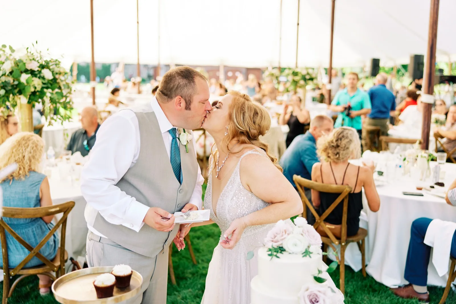 The bride and groom share a kiss beside their tiered wedding cake under a sailcloth tent as guests mingle at tables in the background. — Tim Larsen Photography, Brainerd Lakes MN