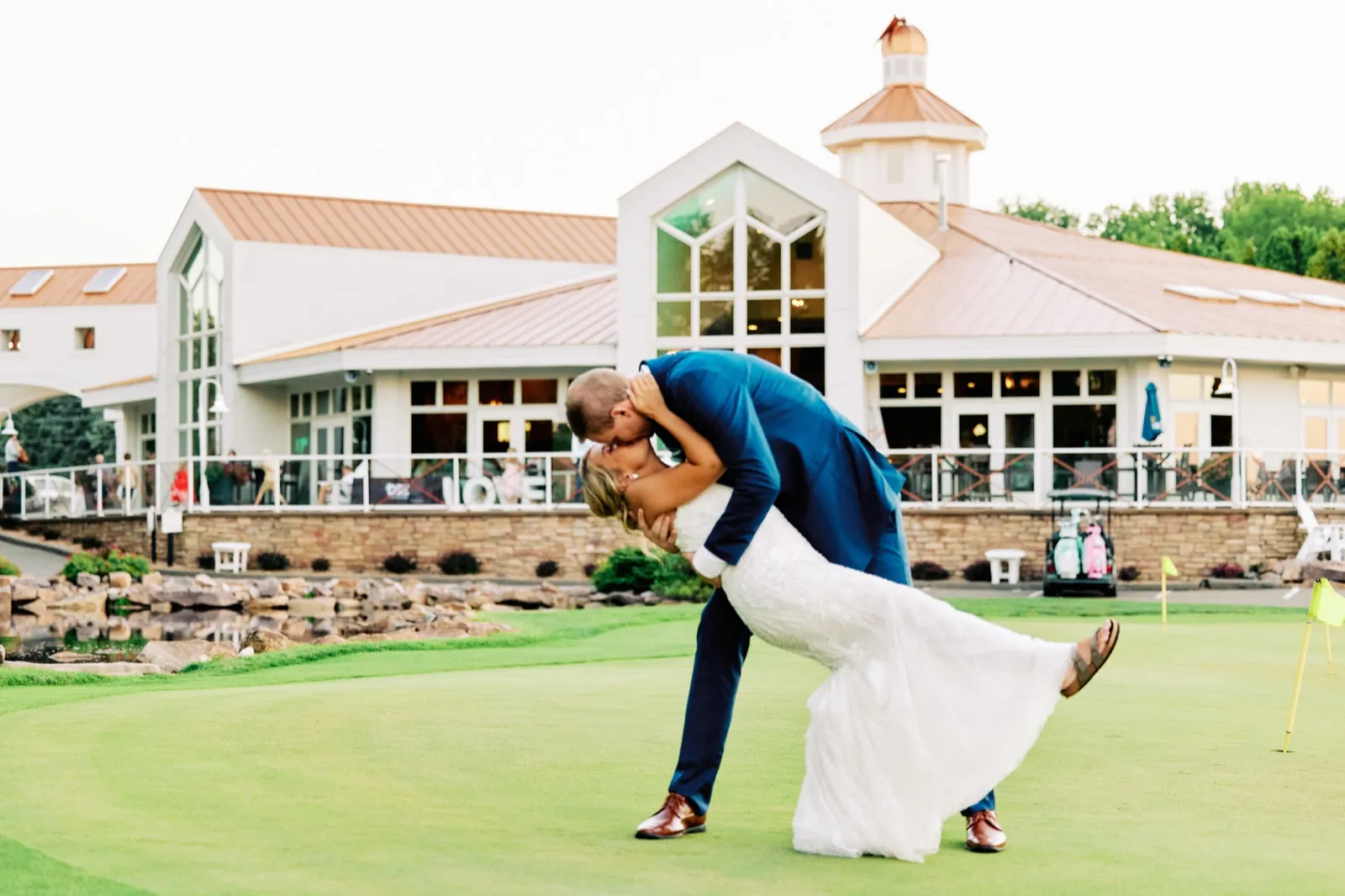 The groom dips the bride into a kiss on a golf course putting green with the Stonebrooke clubhouse and cupola rising behind them. — Tim Larsen Photography, Twin Cities MN