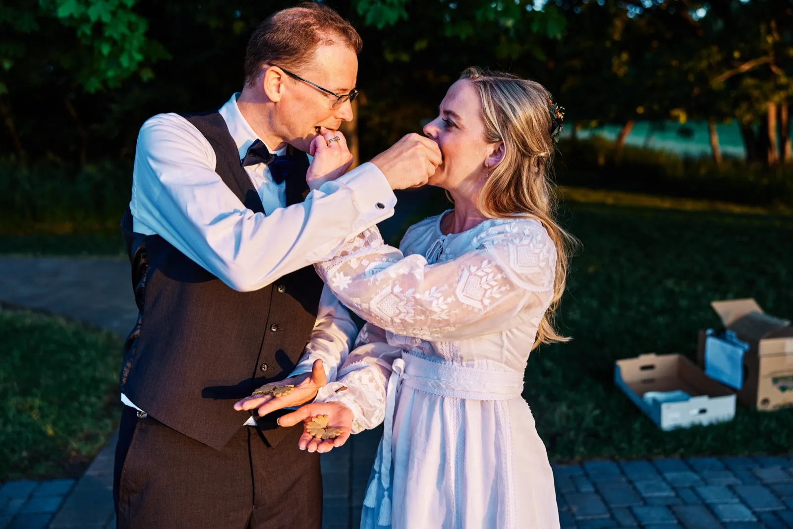 The bride and groom feed each other cookies in warm evening light along a garden path, both mid-bite with hands at each other's faces. — Tim Larsen Photography, Minnesota