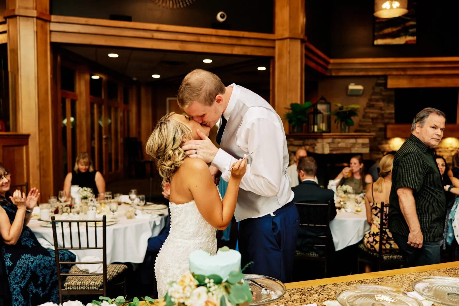 The bride and groom kiss above their small tiered cake while seated guests look on in a warmly lit wood-paneled reception room. — Tim Larsen Photography, Twin Cities MN