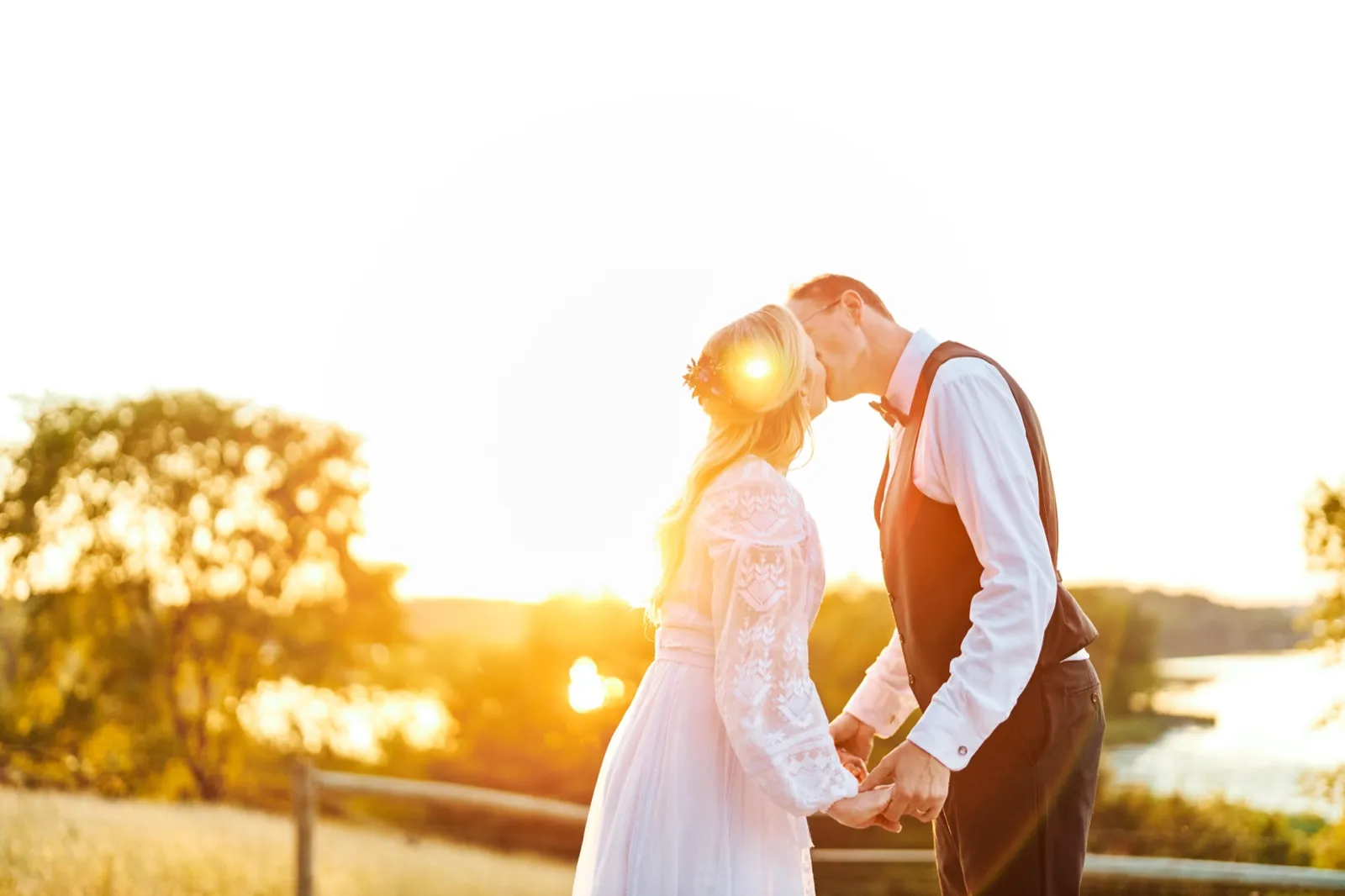 The bride and groom kiss on a hilltop above a lake while the setting sun flares brightly between them. — Tim Larsen Photography, Minnesota