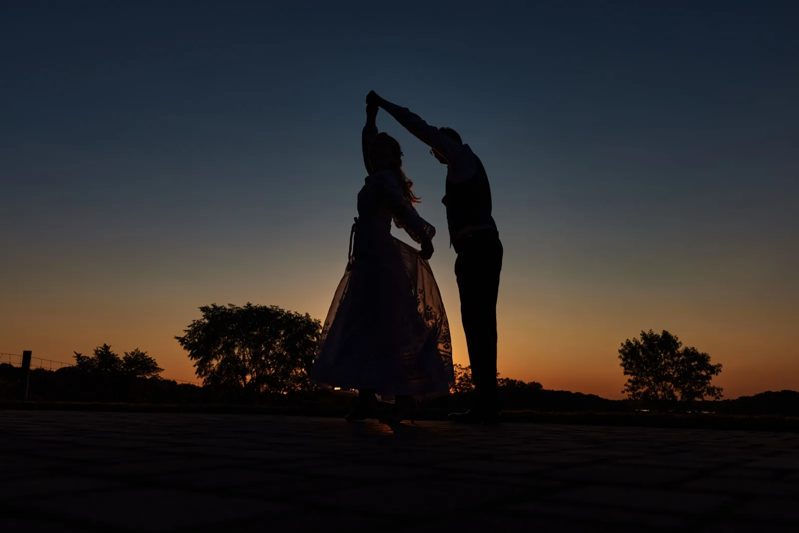 The couple is silhouetted against a deep blue and orange dusk sky as the groom twirls the bride, her dress flaring at her side. — Tim Larsen Photography, Minnesota