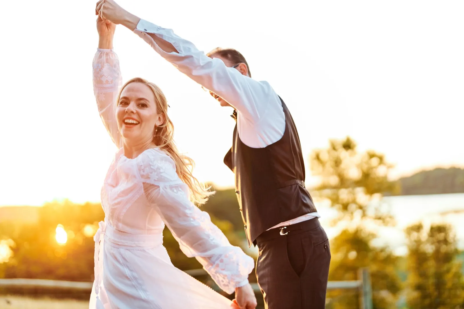 The groom twirls the bride in a lakeside field at sunset, her embroidered white dress catching the warm light as she laughs toward the camera. — Tim Larsen Photography, Minnesota