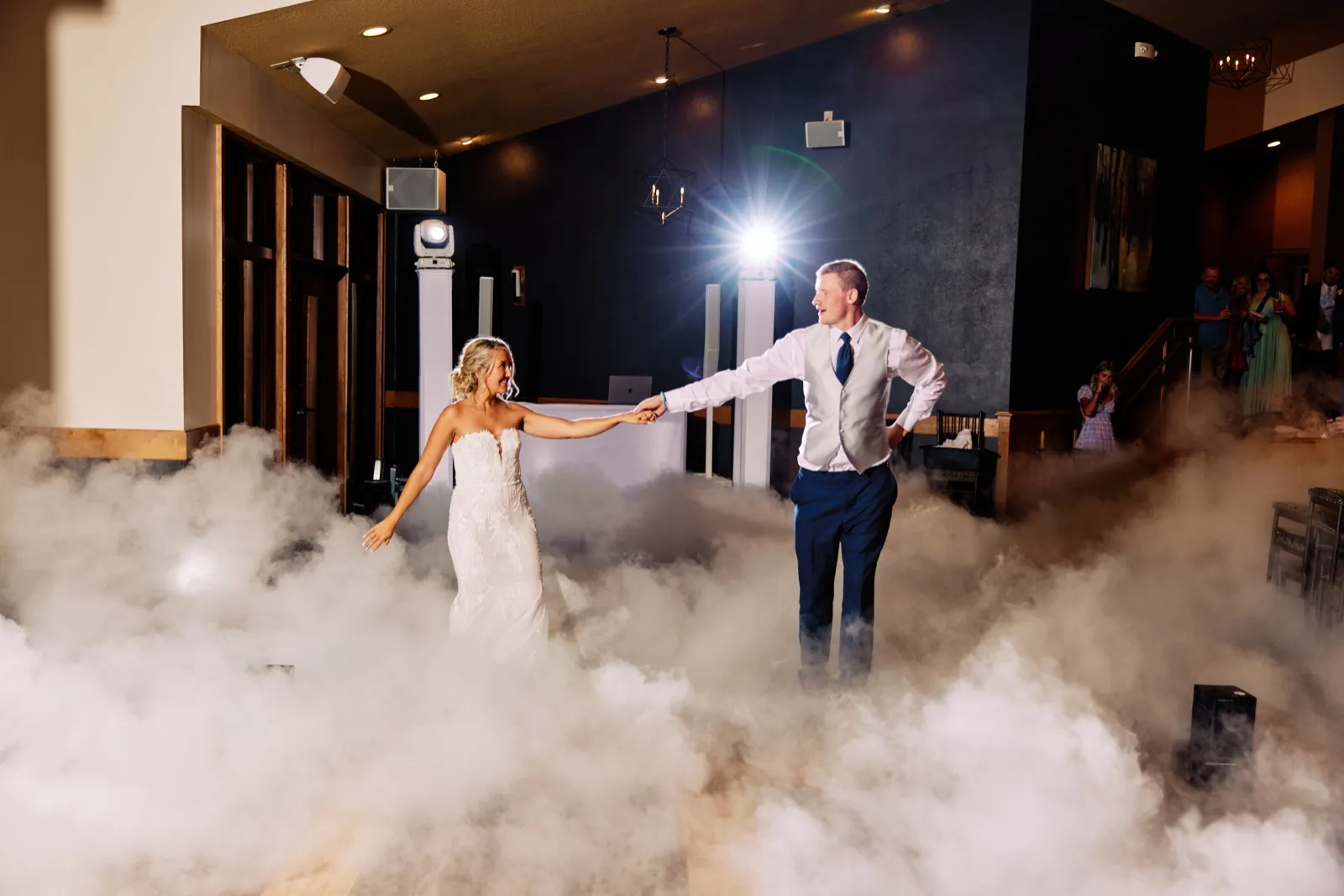 The bride and groom hold hands on a fog-covered dance floor as a stage light flares behind them and guests watch from a staircase. — Tim Larsen Photography, Twin Cities MN