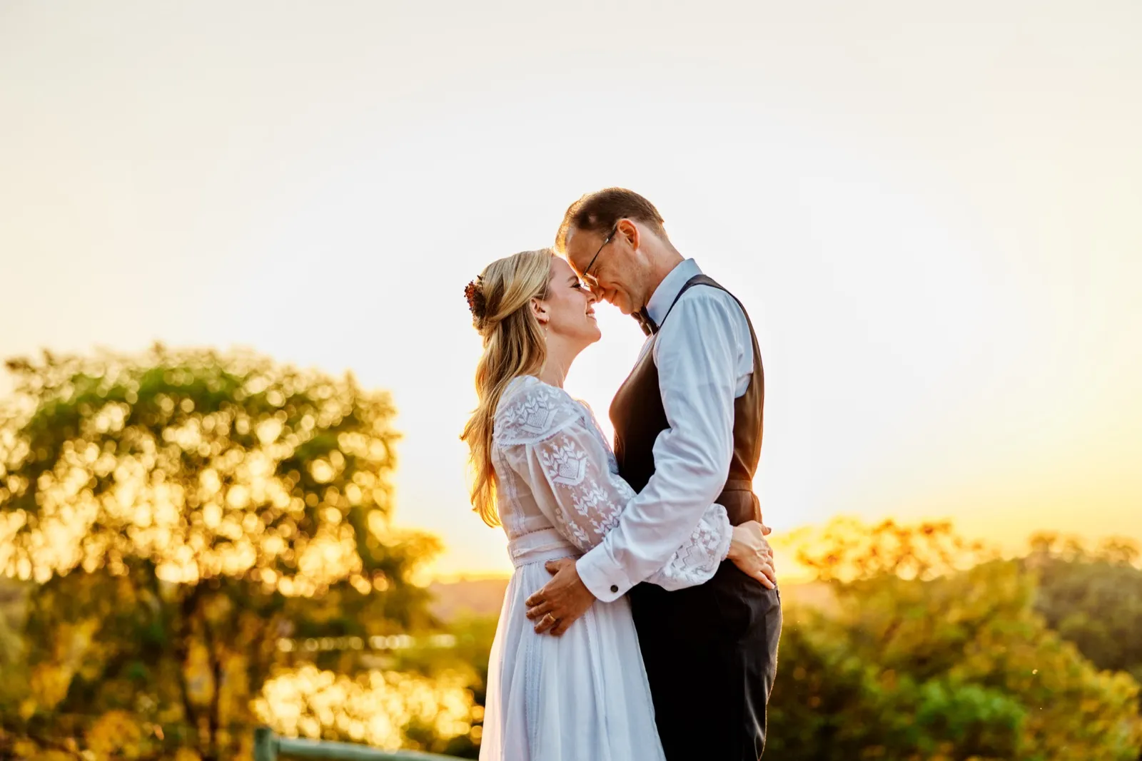 The bride and groom stand forehead to forehead on a hilltop, backlit by a golden sunset over distant trees. — Tim Larsen Photography, Minnesota