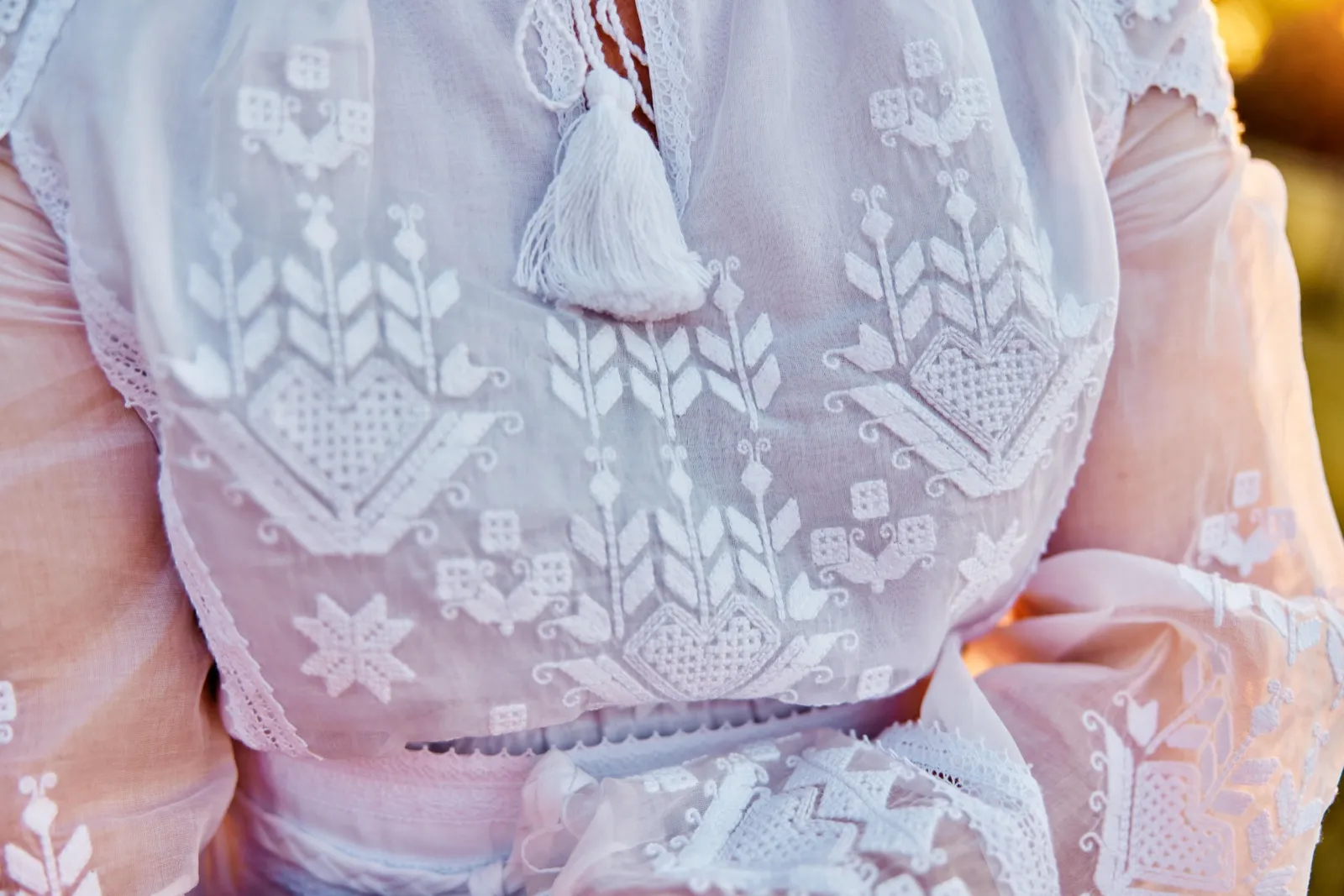 An extreme close-up of the bride's white embroidered bodice with a delicate hanging tassel, lit by warm golden-hour light. — Tim Larsen Photography, Minnesota